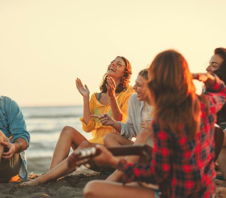 Happy friends sitting on the beach singing and playing guitar during the sunset